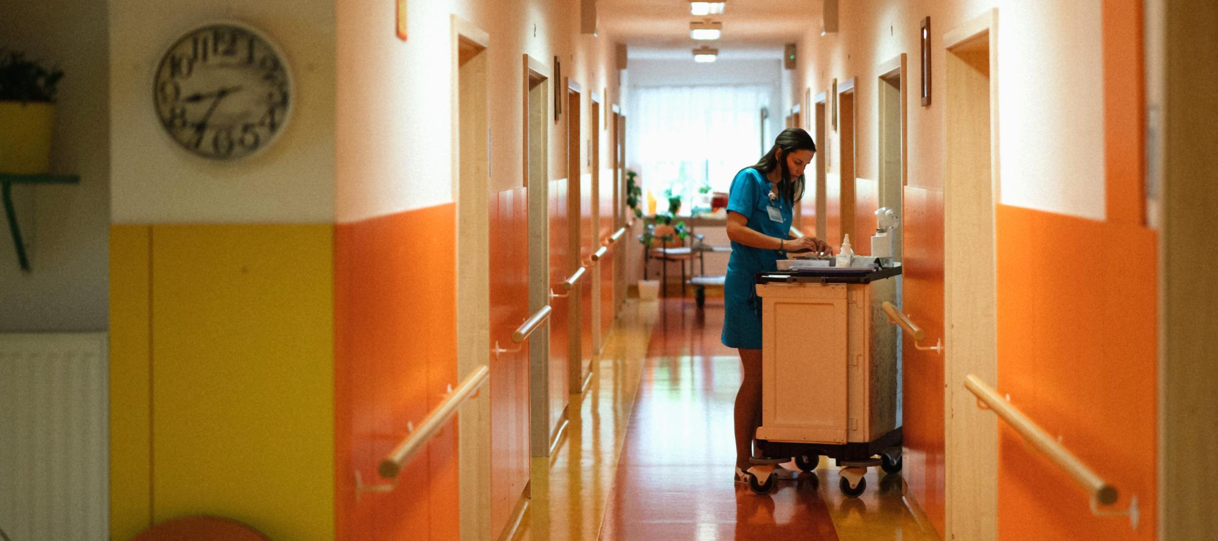 A nurse in a teal uniform works at a medicine cart halfway down a bright, orange-walled residential care corridor.